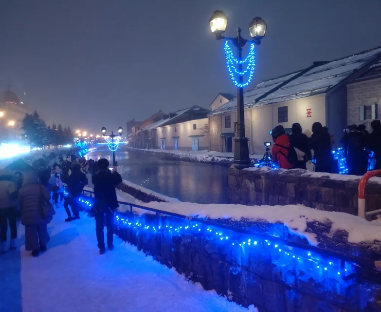 The Otaru Canal at night, lined with blue lights and old street lamps, with people walking along the snowy promenade beside the warehouses.
