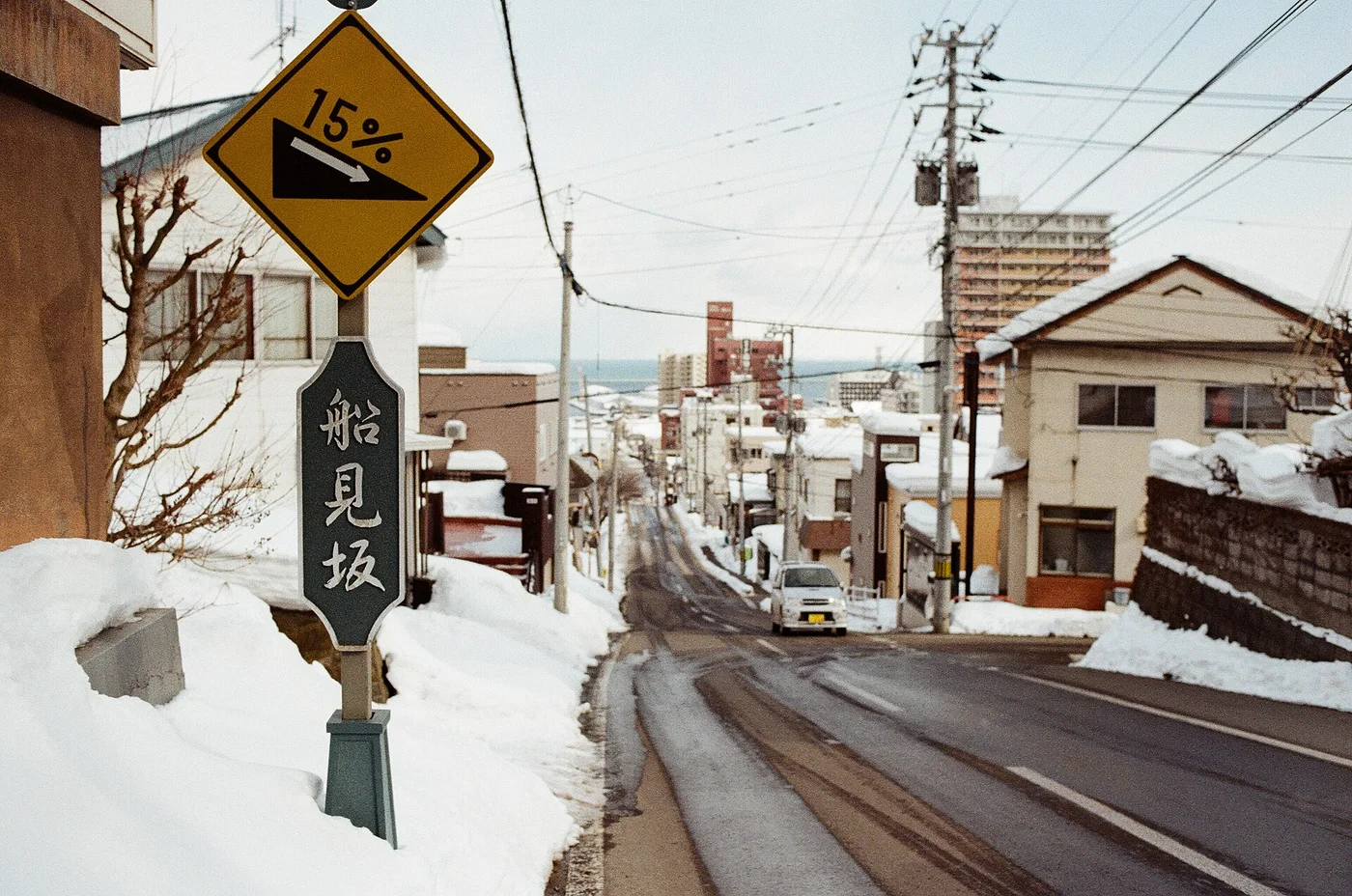 A snowy street in Otaru running steeply downhill towards the sea, with a yellow 15% gradient sign and a sign reading Funami-zaka, snowbanks on both sides.