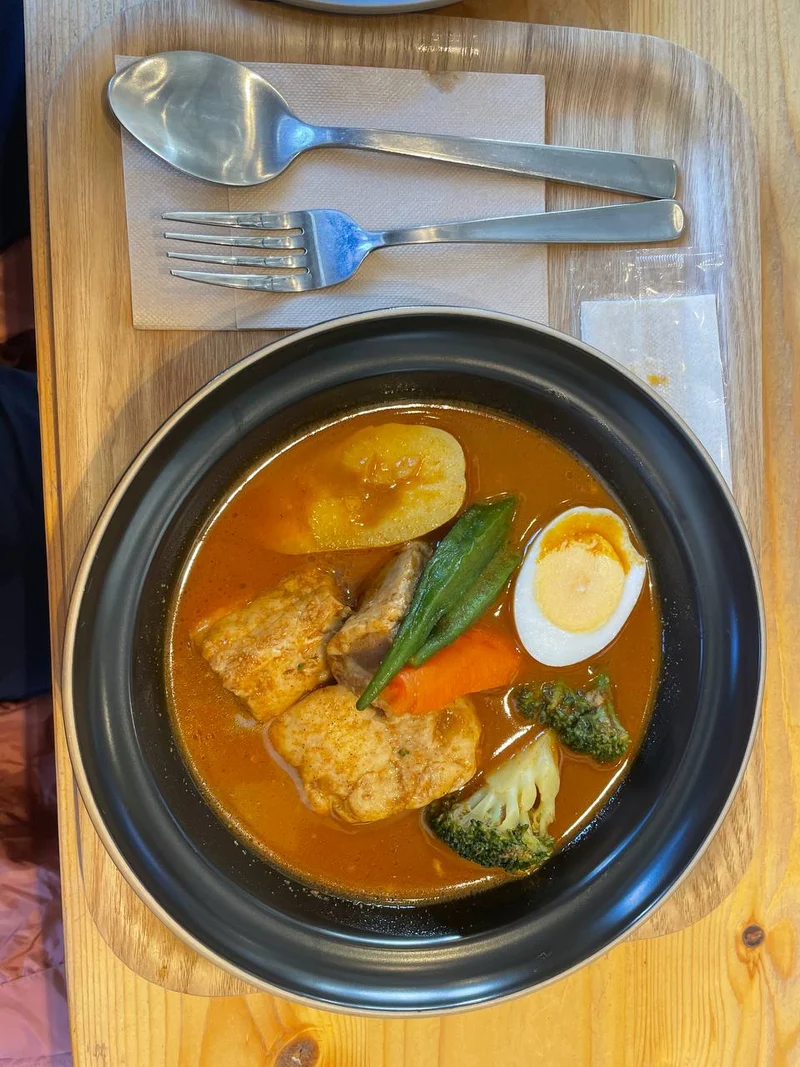 A bowl of Hokkaido soup curry with fried chicken, half a boiled egg, potato, carrot, broccoli and green pepper, on a wooden tray.