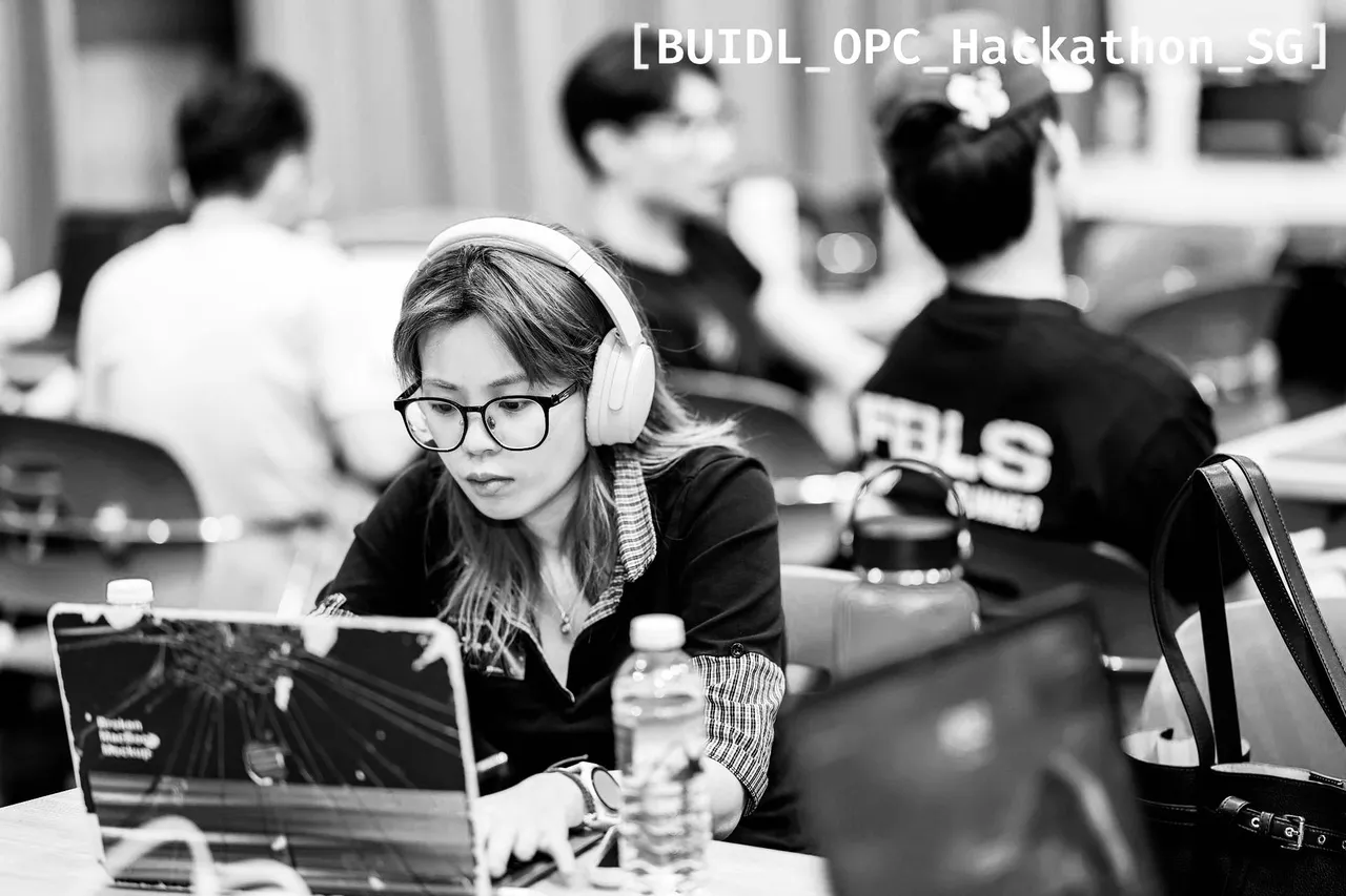 Martina at a hackathon table, headphones on, typing on a laptop, with other builders out of focus behind her.